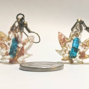May include: A pair of gold and blue resin leaf earrings with a clear background. The earrings are hanging from a silver hook.