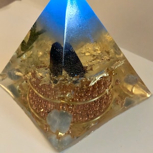 Könnte beinhalten: Eine klare Pyramide mit blauer Spitze und goldenen Akzenten. Im Inneren der Pyramide befinden sich Kupferperlen, Goldflocken und ein schwarzer Kristall.