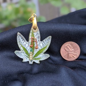 May include: A gold pendant shaped like a marijuana leaf with a green and white design. A musical note is embedded in the leaf. The pendant is about the size of a US penny.