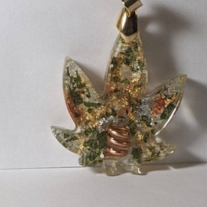 May include: A gold pendant in the shape of a marijuana leaf with green, gold, and copper accents. The pendant is made of resin and has a clear, glassy finish.
