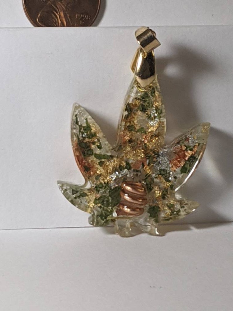 May include: A gold pendant in the shape of a marijuana leaf with green, gold, and copper accents. The pendant is made of resin and has a clear, glassy finish.