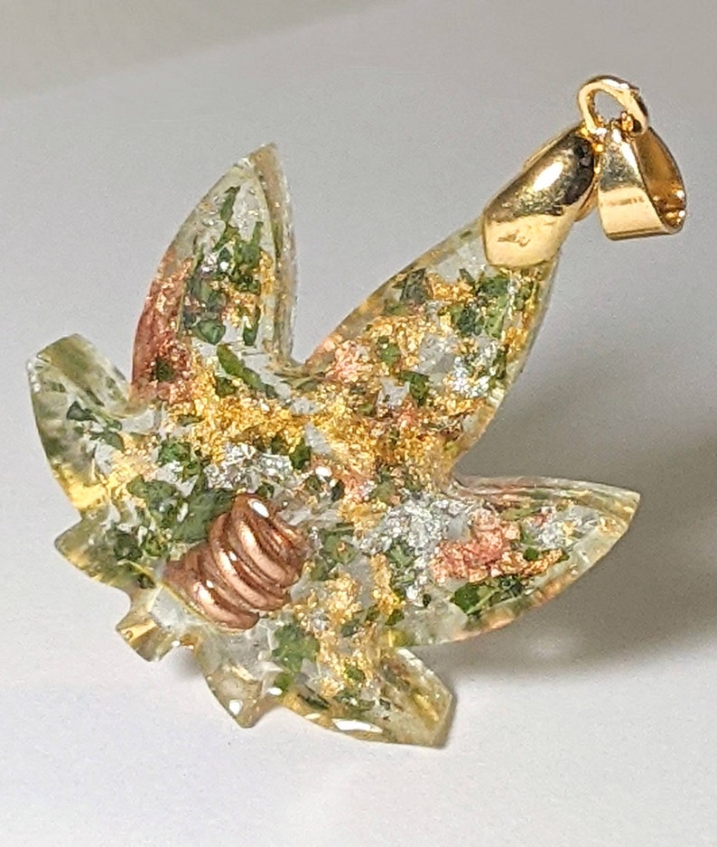 May include: A gold-tone pendant in the shape of a marijuana leaf with green, gold, and silver glitter embedded in clear resin. The pendant has a copper-tone bail.
