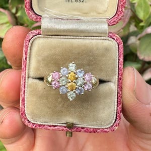 Op de afbeelding: Een gouden ring met een cluster van kleurrijke edelstenen, waaronder roze, gele en blauwe stenen, gepresenteerd in een vintage roze sieradendoosje. Het doosje is open en onthult de ring tegen een beige fluwelen interieur.