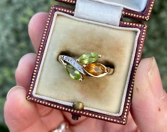 May include: A gold ring with a leaf design, featuring three gemstones in shades of green and orange. A section of the ring is adorned with small, clear stones. The ring is displayed in a vintage jewelry box with a beige interior.