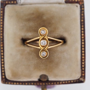 May include: A gold ring with three small diamonds set in a unique, swirling design. The ring is displayed in a vintage jewelry box with a soft, beige interior. The ring's design is symmetrical and elegant.