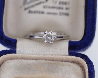 Puede incluir: Un anillo de compromiso de plata con un solo diamante redondo en una caja de terciopelo azul.
