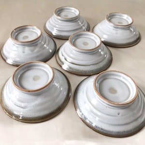 Może przedstawiać: Zestaw sześciu białych ceramicznych misek z brązową obw&oacute;dką. Miski mają lekko fakturowaną powierzchnię i są ułożone jedna na drugiej.