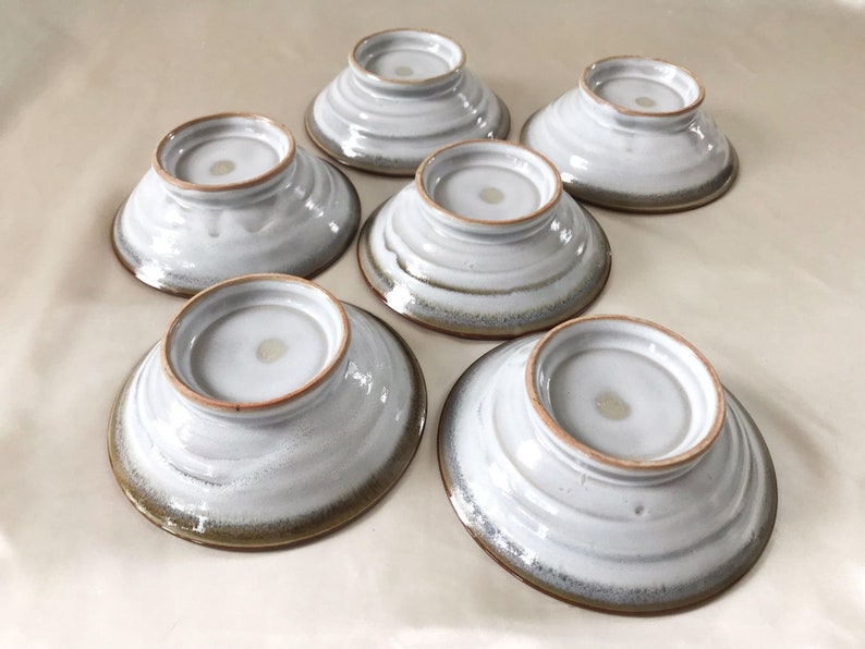 Może przedstawiać: Zestaw sześciu białych ceramicznych misek z brązową obw&oacute;dką. Miski mają lekko fakturowaną powierzchnię i są ułożone jedna na drugiej.