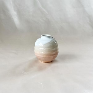 May include: A small, round, ceramic vase with a white, beige, and pink glaze. The vase has a textured surface and a slightly rounded opening.