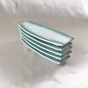 May include: A stack of five light green rectangular ceramic plates with a gold rim. The plates are stacked on a white surface.