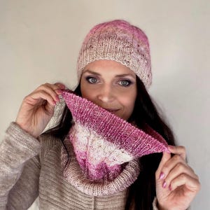 May include: A pink and white knitted hat and matching cowl. The hat features a cable knit pattern and the cowl has a lacy design.