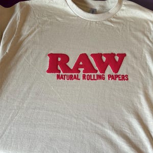 Op de afbeelding: Een beige T-shirt met een rood RAW-logo en de tekst "NATURAL ROLLING PAPERS" gedrukt op de voorkant.