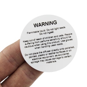 May include: A white round warning label with black text. The label reads "WARNING Flammable liquid. Do not light reeds. Do not ingest. Keep out of reach of children and pets. People suffering from perfume sensitivity should be cautious when using this product. Use gloves when handling used reeds. Do not place the diffuser directly onto polished, painted, plastic or leather surfaces or on electrical equipment, as accidental spillage may cause damage to certain materials."