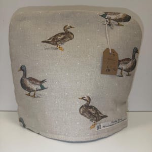 May include: A beige fabric tea cozy featuring a repeating pattern of brown and green ducks with small white polka dots. A brown rectangular paper tag is attached with string, and a small white fabric label reads Verity Rose Handmade.