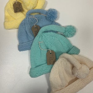 Può includere: Quattro cappellini di lana per neonati con pon pon di colore giallo, blu, verde e crema. Ogni cappellino ha un'etichetta che dice "Verity & Rose".