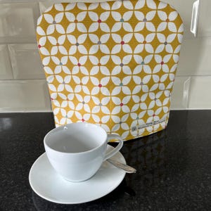 Könnte beinhalten: Ein Teewärmer mit gelbem Blumenmuster, eine weiße Teetasse und eine Untertasse. Der Teewärmer hat ein sich wiederholendes weißes Blumendesign mit roten, blauen und grauen Akzenten. Ein silberner Löffel liegt auf der Untertasse.