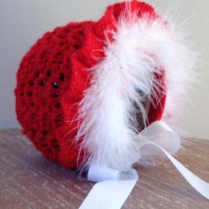 May include: A red crocheted baby bonnet with white feather trim and a white satin ribbon. The bonnet is a festive holiday accessory, perfect for a baby's first Christmas or winter photos.