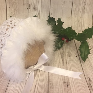 May include: White crocheted baby bonnet with a white faux fur trim and a white satin ribbon.