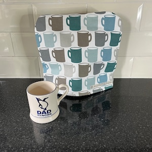 May include: A white mug with a blue penguin graphic and the words "DAD DANCING" sits in front of a patterned appliance cover. The cover features a repeating pattern of mugs in shades of blue, gray, and brown on a white background.