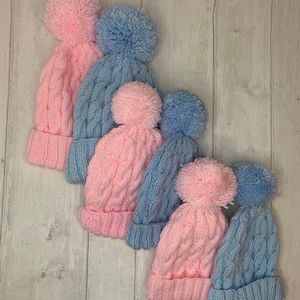 Puede incluir: Cinco gorros de punto con pompones en colores rosa y azul alternados. Los gorros están dispuestos en fila, con el gorro más grande en la parte superior y el más pequeño en la parte inferior.