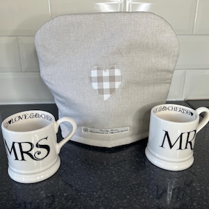 May include: A beige tea cozy with a heart-shaped, checkered patch, and two white mugs. One mug says "MRS" and the other says "MR" in black script. The mugs have the words "LOVE & CHERISH" printed on the rim.