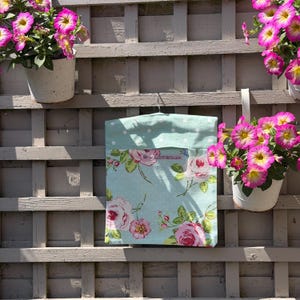 May include: A light blue fabric bag featuring a white polka dot pattern on the top section and a floral print with pink roses and green leaves on the lower section. The bag is suspended from a metal hanger against a wooden lattice wall.