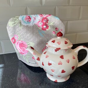 May include: A white teapot with a red heart pattern and a gray floral tea cozy. The tea cozy has a white polka dot pattern and the text "Handmade" is visible on the bottom.
