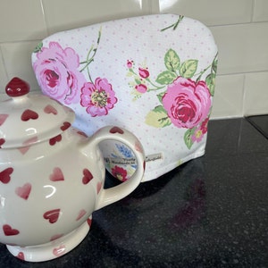 Può includere: Una teiera bianca con un motivo a forma di cuore rosa e rosso. La teiera è appoggiata su un piano di lavoro nero accanto a un copriteiera bianco con un motivo floreale rosa. Il copriteiera ha un'etichetta che dice "Verity Handmade in England" e "Wellerfield".