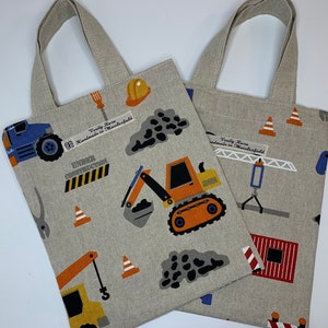 May include: Two beige tote bags with construction vehicle patterns. The bags feature diggers, cranes, and safety cones. The words "UNDER CONSTRUCTION" are also visible. Each bag has a matching beige handle.