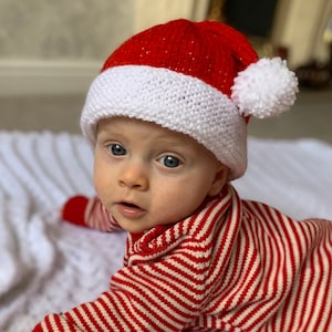 Peut inclure: Un bébé portant un bonnet de Noël rouge et blanc avec un pompon blanc sur le dessus. Le bébé porte un pull rayé rouge et blanc.