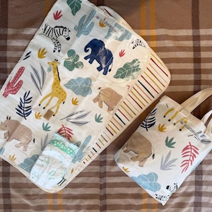 May include: A white baby changing mat and matching tote bag featuring a safari animal print. The print includes a giraffe, zebra, elephant, and rhino, with colourful leaves and cacti. A diaper is also visible.