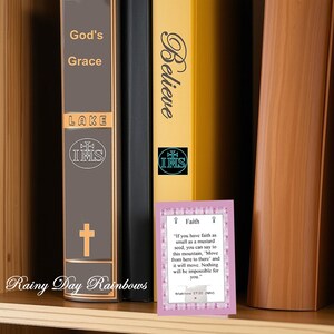 May include: A wooden bookshelf displaying three books and a small card. The books are titled "God's Grace", "Believe", and "LAKE". The card features a quote about faith from Matthew 17:20. The text "Rainy Day Rainbows" is visible.