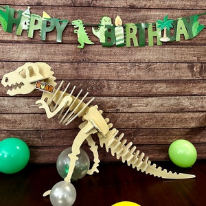 May include: A wooden dinosaur skeleton cutout with the word "ROAR" on its neck. The dinosaur is standing on a wooden floor with green and silver balloons. A green banner with the words "Happy Birthday" is hanging on a brown wooden wall.