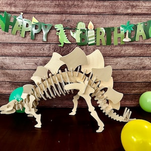 May include: A wooden dinosaur skeleton with a green and brown 'Happy Birthday' banner hanging above it. There are green and yellow balloons on the floor.