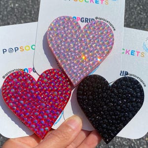 May include: Three heart-shaped phone grips on white backing cards labeled POPSOCKETS. One grip is covered in iridescent light pink rhinestones, one in iridescent red rhinestones, and one in solid black rhinestones. Each card features the text @aplacetopixie.