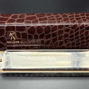 May include: A silver Hohner Chromonica 280-C harmonica with a brown faux-leather case. The harmonica has 12 holes and the text "Hohner" and "Made in Germany" are visible. The case has a reptile skin pattern.