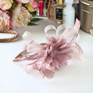 May include: A blush pink headband featuring a large flower and feathers. The headband is made from a soft, delicate material and is ideal for adding a touch of elegance to any outfit.