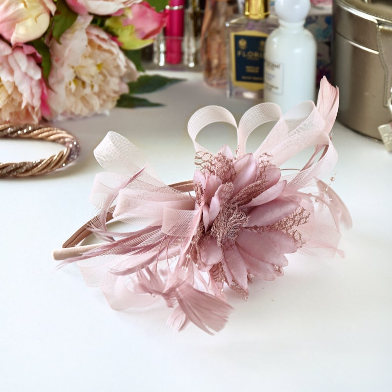 May include: A dusty rose headband featuring a large fabric flower centerpiece, layered sheer mesh loops, and delicate feathers. The accessory is accented with fine, thin stems and small beads, creating a textured, dimensional floral design.