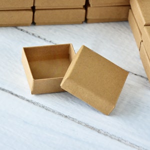 May include: An open, square, cardboard box with a separate lid. The box is a light brown colour and is on a white and grey wooden surface. Several other boxes are visible in the background.