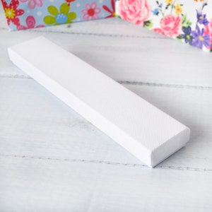 May include: A white rectangular box with a textured surface. The box is lying on a white wooden surface.