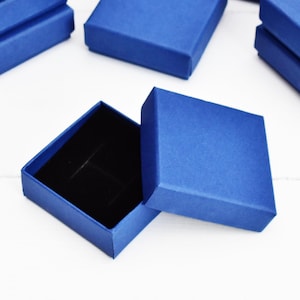 May include: A collection of small, square, royal blue gift boxes, one open to show a black velvet interior. The boxes are on a white surface. These are likely for jewellery or small presents.