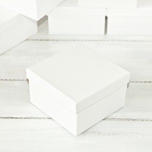 May include: A white cardboard box with a textured surface. The box is empty and sitting on a white wooden surface.