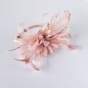 May include: A dusty rose fascinator headband featuring a central fabric flower, delicate mesh loops, and thin feathers. The accessory is constructed on a slim, matching satin-covered band, creating a layered, textured appearance in a soft pink hue.