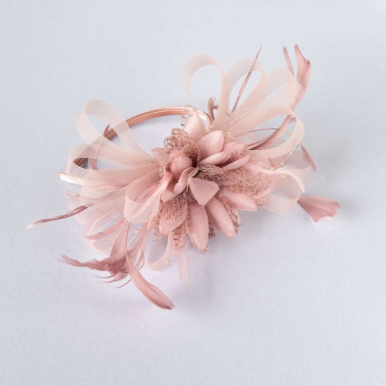 May include: A dusty rose fascinator headband featuring a central fabric flower, delicate mesh loops, and thin feathers. The accessory is constructed on a slim, matching satin-covered band, creating a layered, textured appearance in a soft pink hue.