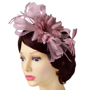 May include: A dusty rose fascinator headpiece featuring a large fabric flower, delicate lace detailing, sheer mesh loops, and thin feathers. The accessory is adorned with small white beads and is designed to be worn on the side of the head.