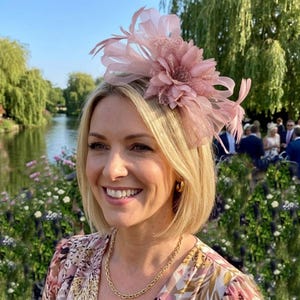 May include: A dusty pink fascinator headpiece featuring a large fabric flower, delicate mesh petals, and thin, wispy feathers. The accessory is designed with a layered, dimensional structure that sits atop the head, showcasing a soft, monochromatic color palette.