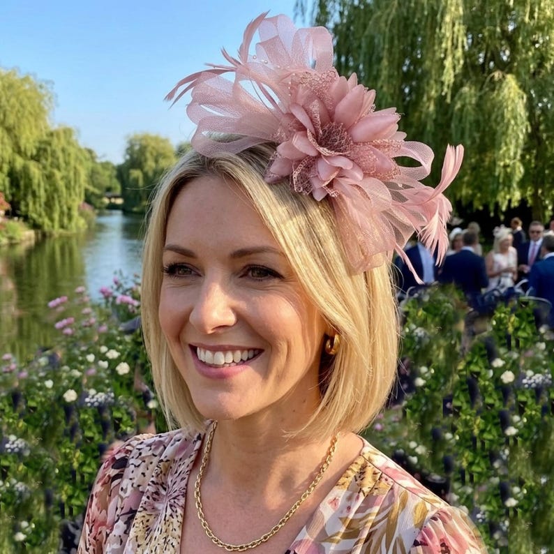 May include: A dusty pink fascinator headpiece featuring a large fabric flower, delicate mesh petals, and thin, wispy feathers. The accessory is designed with a layered, dimensional structure that sits atop the head, showcasing a soft, monochromatic color palette.