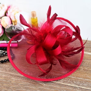 May include: A burgundy fascinator with a large, curled bow and feathers. The fascinator is made of a fine mesh fabric and has a wired edge.