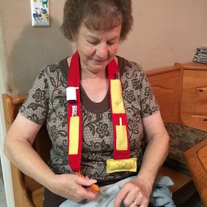 Puede incluir: Una mujer lleva un cordón de tela rojo y amarillo con bolsillos y un pequeño contenedor adjunto. Está sosteniendo un par de tijeras y está sentada en un banco de madera.