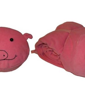 Puede incluir: Una manta de forro polar rosa con forma de cerdo con ojos negros y un hocico. La manta se puede enrollar en forma de cerdo.